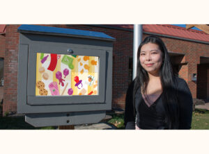 Jessica Cheng stands beside her mini gallery, displaying a colourful artwork featuring illustrations of food.