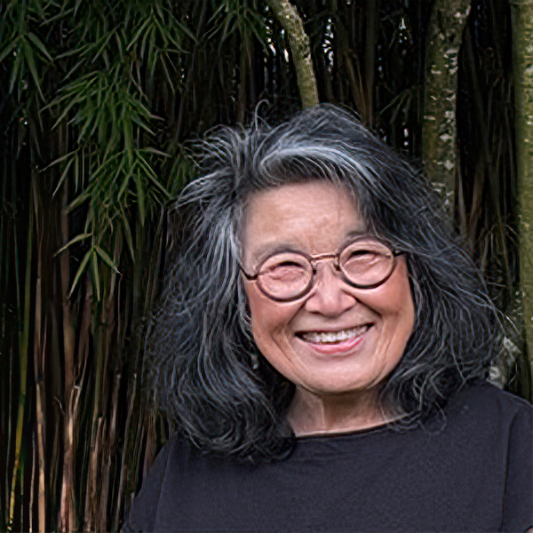 Marcia Iwasaki smiling with gray, wavy hair, round glasses, wearing a dark top with a dark nature background.