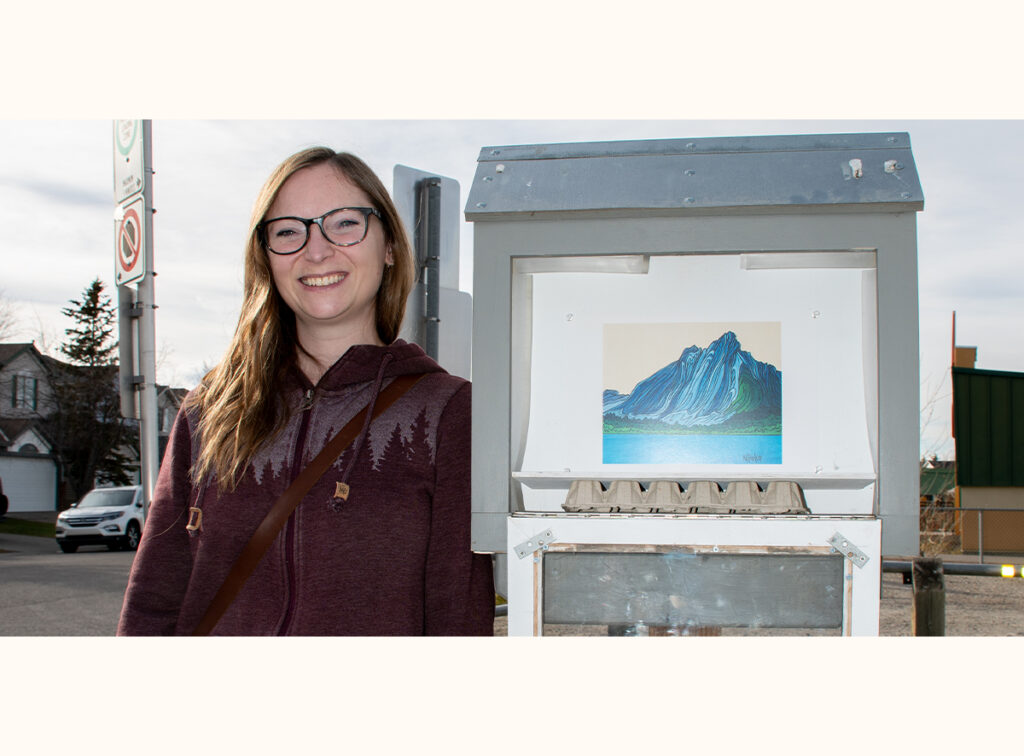 Megan Parker stands beside her mini gallery, displaying a landscape artwork of a mountain by a lake.