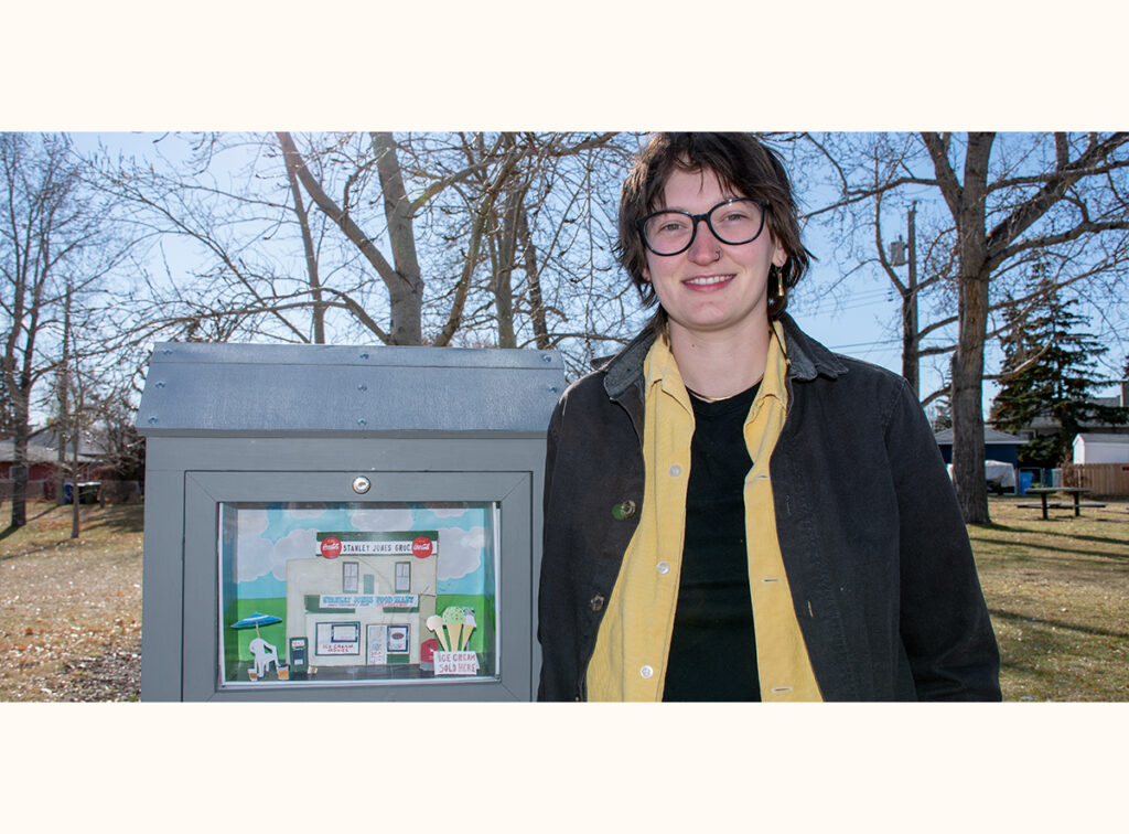 Rachel Kolodychuk stands beside their mini gallery, which displays artwork of a small grocery store scene.