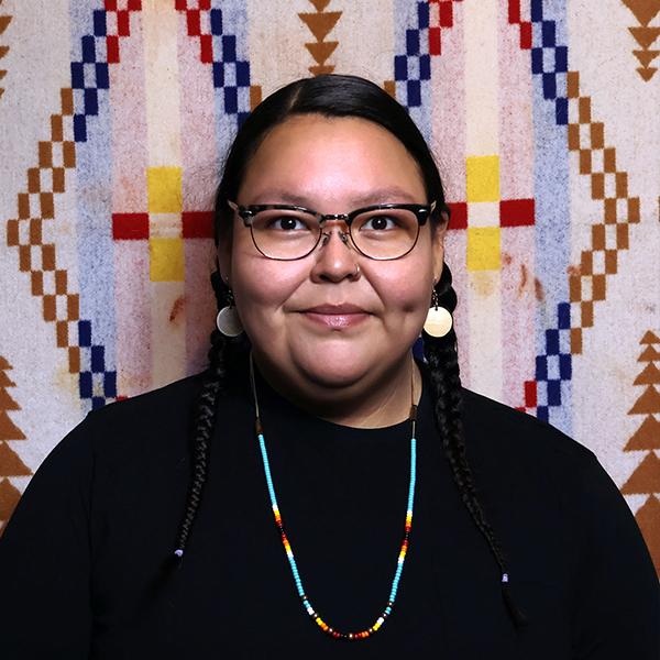 Sikapinakii Low Horn with glasses, braids, a black shirt and a beaded necklace.