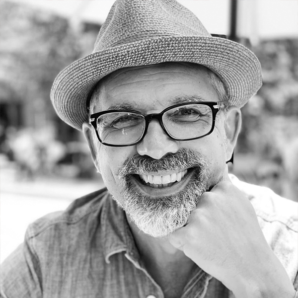 Stephen Duncombe in a black-and-white portrait, wearing glasses and a hat, smiling, with a bearded chin resting on their hand.