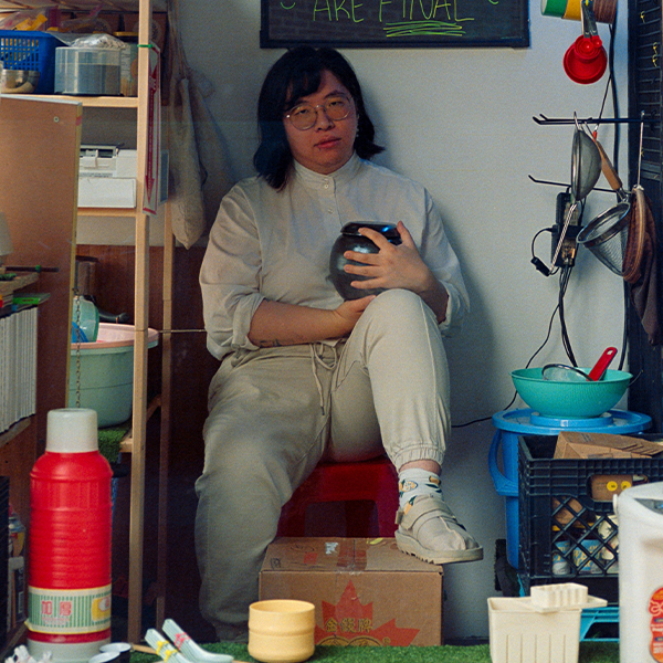 Teresa Tam wearing all white and glasses sits in a small space filled with objects, with a foot propped on a box.