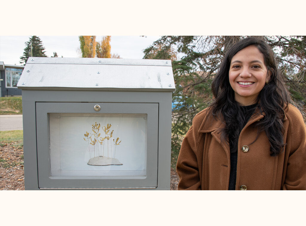 Victoria Sanchez stands beside her mini gallery, featuring artwork of golden plant stems on a small mound.