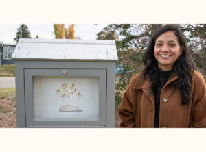 Victoria Sanchez stands beside her mini gallery, featuring artwork of golden plant stems on a small mound.