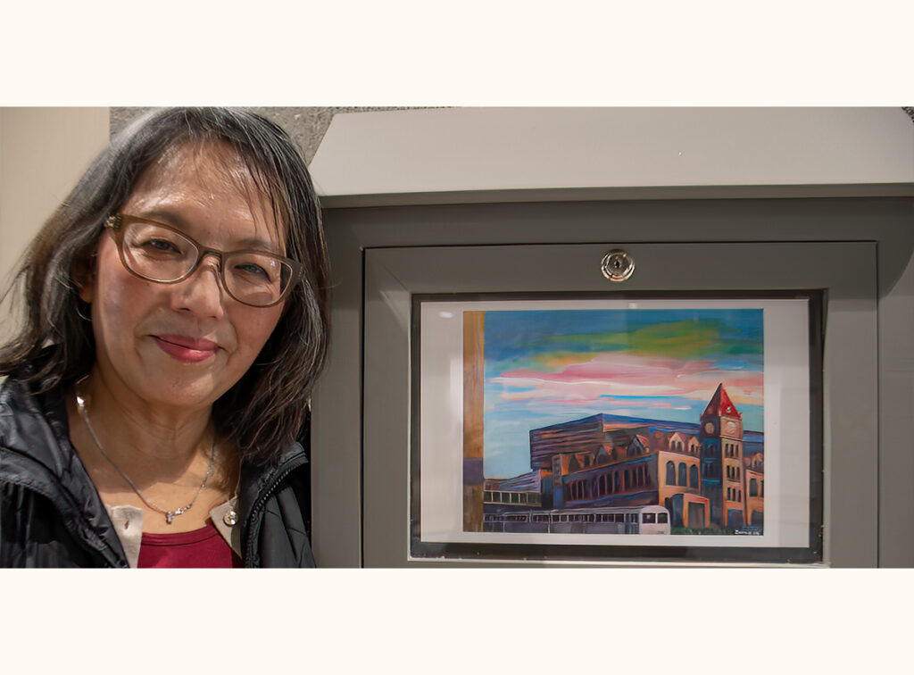 Artist Zoong Nguyen standing beside her artwork of a vibrant cityscape of Calgary city hall at sunset.