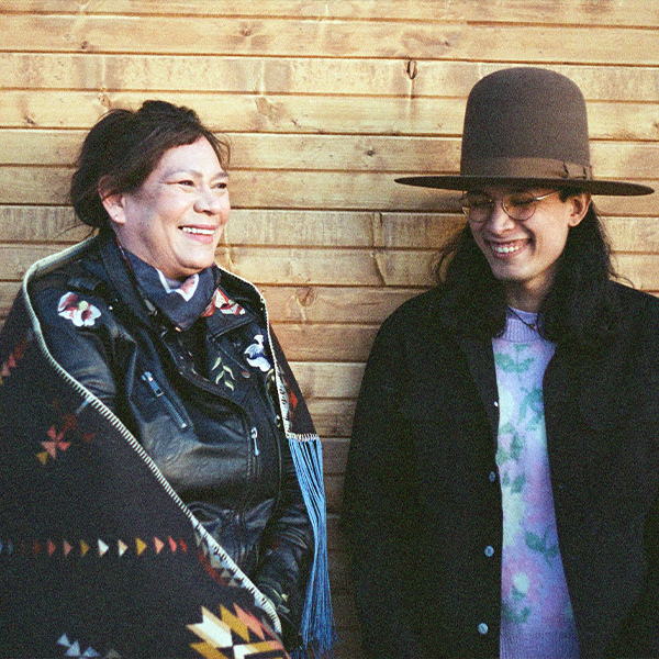 tīná gúyáńí artist collective's Glenna Cardinal smiling and wearing an embroidered leather jacket and scarf. Next to Glenna, seth cardinal dodginghorse, smiling with glasses and eyes closed, wearing a brown hat, colourful shirt and black jacket.  