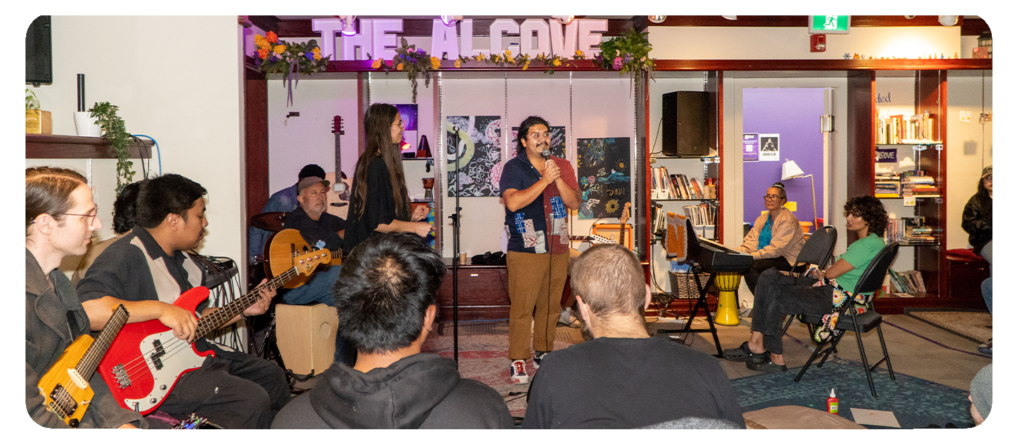 An indoor event with people gathered around a stage, listening to a speaker with musicians seated.