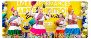 A group of women perform a traditional cultural dance in front of a bright yellow backdrop that reads “Live Performances & Art Exhibition Extravaganza.” They wear colourful skirts with embroidered patterns, white lace tops and woven sashes, each holding a vibrant hat. The stage is decorated with blue, gold, and white balloons and logos for Calgary Arts Development, Canada Zalmi Society and ILMI are visible in the background.