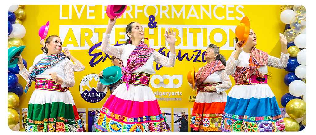 A group of women perform a traditional cultural dance in front of a bright yellow backdrop that reads “Live Performances & Art Exhibition Extravaganza.” They wear colourful skirts with embroidered patterns, white lace tops and woven sashes, each holding a vibrant hat. The stage is decorated with blue, gold, and white balloons and logos for Calgary Arts Development, Canada Zalmi Society and ILMI are visible in the background.