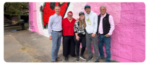 Five people stand smiling in front of a bright pink mural wall featuring a large red and black illustration. They are posed closely together on a paved surface, with trees and a black mailbox visible in the background.