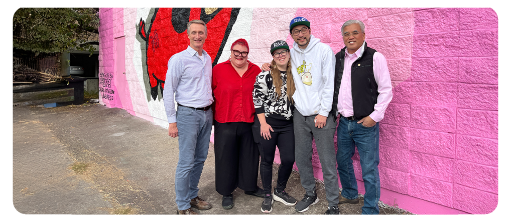 Five people stand smiling in front of a bright pink mural wall featuring a large red and black illustration. They are posed closely together on a paved surface, with trees and a black mailbox visible in the background.