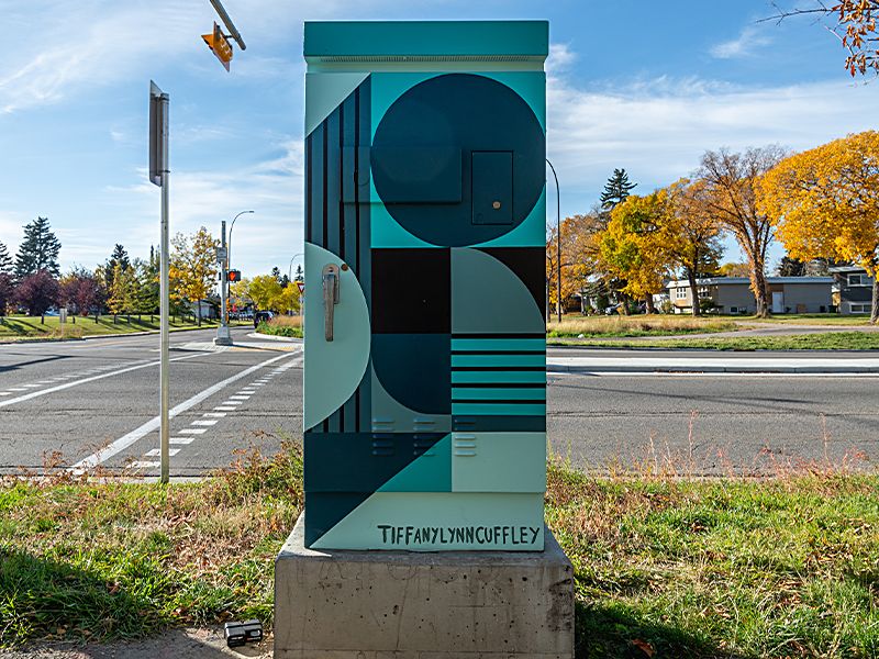 A utility box painted with shapes in shades of blue, teal, and black, with circles, stripes, and rectangles. The artist's name, "Tiffany Lynn Cuffley," is at the bottom.