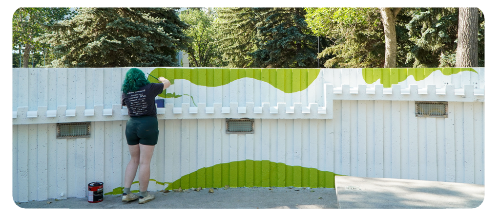 A person with green hair painting a mural with green and white wavy shapes on a wall outdoors.