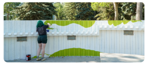 A person with green hair painting a mural with green and white wavy shapes on a wall outdoors.