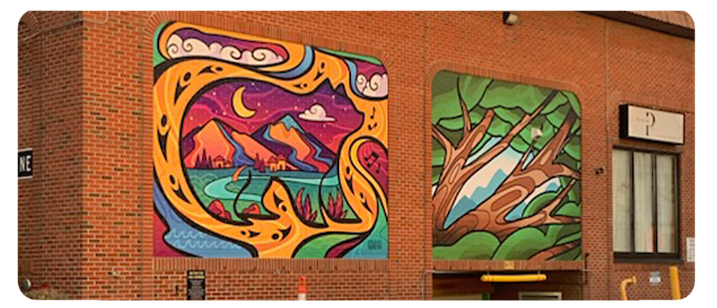 Two murals are painted on the side of a red brick building. The left mural features a mountain landscape with swirling orange shapes, a crescent moon and a purple sky. The right mural depicts green tree branches framing a mountain peak.