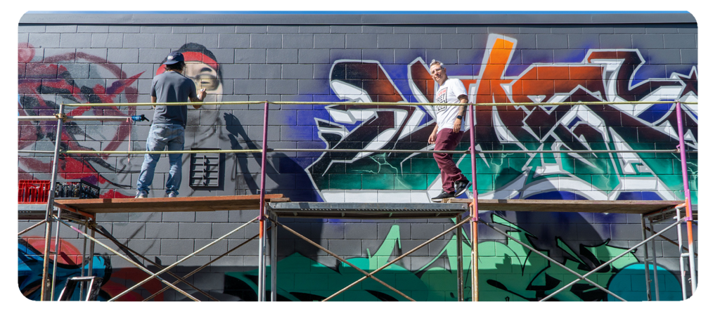 Two people working on a mural while standing on scaffolding, painting graffiti art on a wall.