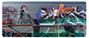 Two people working on a mural while standing on scaffolding, painting graffiti art on a wall.