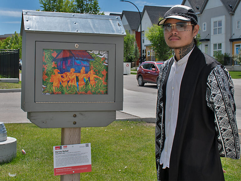 The image shows a man standing beside a mini gallery. The artwork inside the mini gallery depicts a group of people dressed in orange in front of a house surrounded by greenery. The person is wearing a patterned jacket, white shirt, and a hat. There are townhouses and a car in the background.