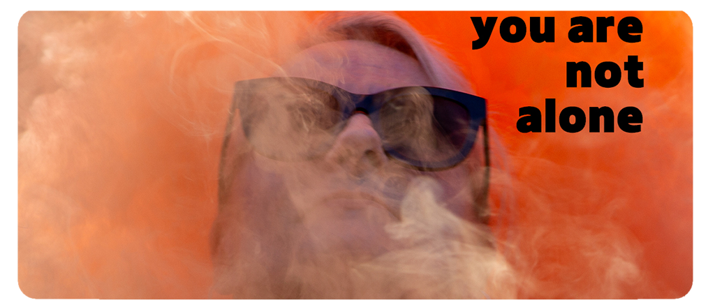 A person wearing large black sunglasses is partially obscured by swirling white smoke. Bold black text on the top right reads, “you are not alone.”