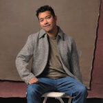 Lionel Migrino sitting on a stool against a brown backdrop wearing a grey jacket