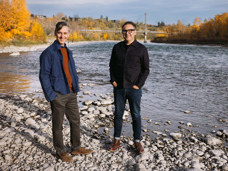 Sans façon artist collective members Charles Blanc (left) & Tristan Surtees (right) stand on the banks of the Bow River, with a pedestrian bridge and trees in the distance.