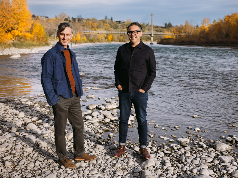 A photograph of Sans façon (Charles Blanc and Tristan Surtees) standing on a rocky riverbank with a river flowing behind them. The background features a bridge and trees with autumn foliage. The man on the left wears a blue jacket and brown pants, while the man on the right is dressed in a black jacket and jeans. Both are smiling at the camera.