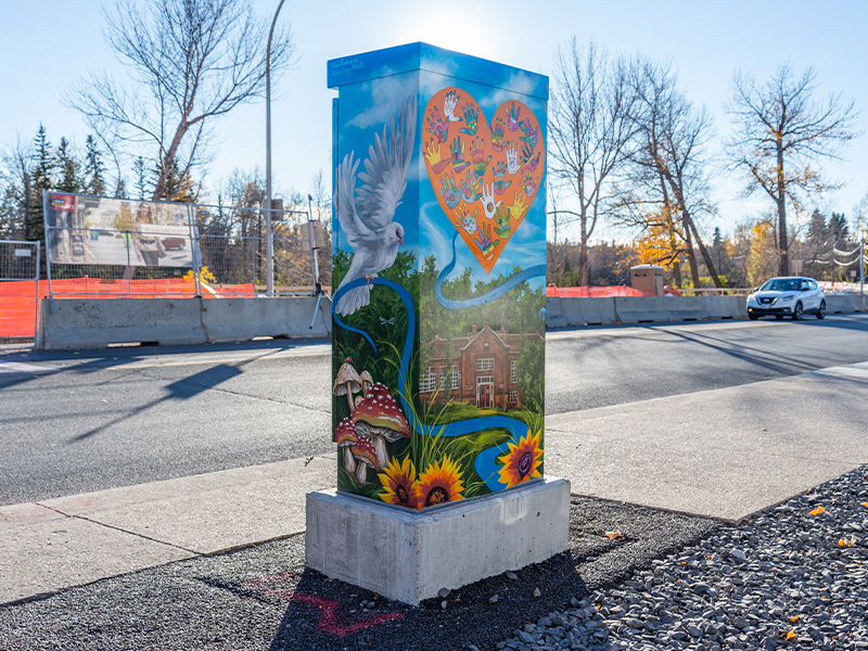 Utility box mural featuring a white dove, heart filled with handprints, mushrooms, sunflowers, and a building in a nature setting.