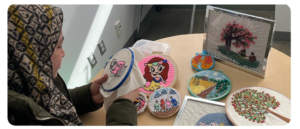 A woman wearing a patterned hijab works on an embroidery hoop featuring a pink cartoon character. Several other colourful embroidery pieces are displayed on a round table, including images of trees, people and landscapes.
