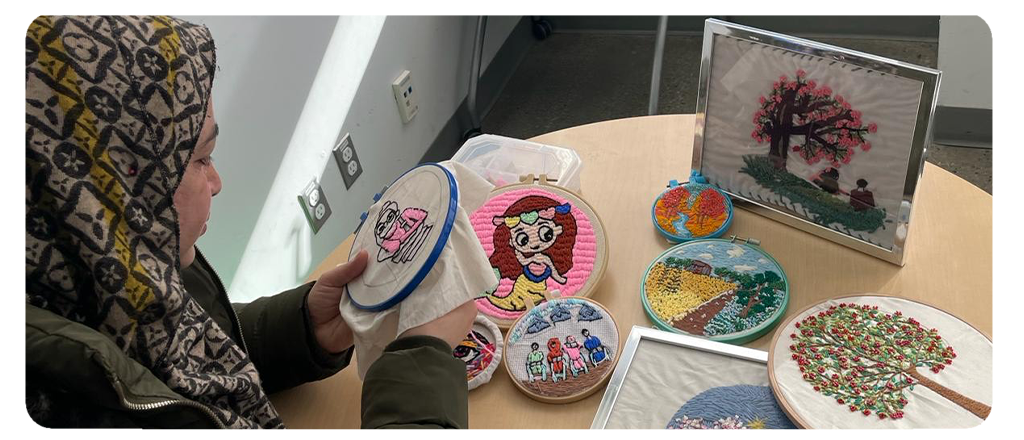 A woman wearing a patterned hijab works on an embroidery hoop featuring a pink cartoon character. Several other colourful embroidery pieces are displayed on a round table, including images of trees, people and landscapes.