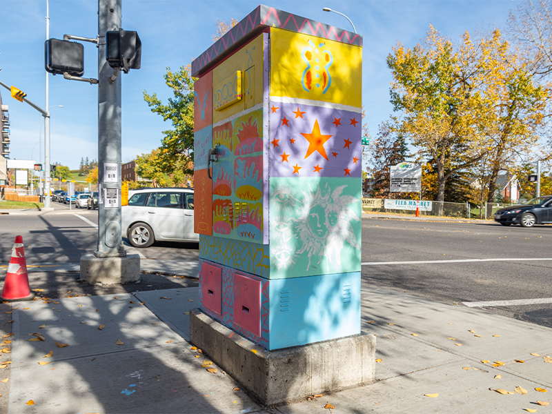 A colourful utility box mural featuring a butterfly, stars, a sun with a face, and various abstract patterns.