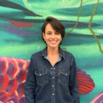 Sayonara Cunha smiling in a denim shirt standing against a mural of a fish in water