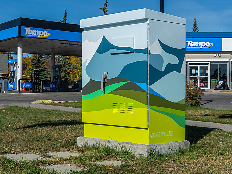 Utility box painted with a landscape featuring mountains in shades of blue and green, against a light sky. The bottom half has green hills.