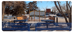 Outdoor seating area with blue picnic tables and wooden benches arranged around a path. Two large signboards with project information are posted at the far end. Snow covers the ground and parked cars are visible in the background.