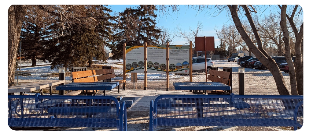 Outdoor seating area with blue picnic tables and wooden benches arranged around a path. Two large signboards with project information are posted at the far end. Snow covers the ground and parked cars are visible in the background.
