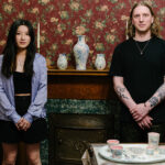 A woman and a man stand side by side in a room with floral wallpaper and a wooden fireplace decorated with blue and white vases. The woman wears a light purple silk button up over a black top and skirt, the man wears a black t-shirt and has tattoos on his arms. A table in the front displays ceramic.
