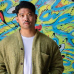 A portrait of a man with short black hair and a moustache, wearing a white t-shirt and a green cardigan. He is standing in front of a graffiti mural with bright colours and abstract shapes.