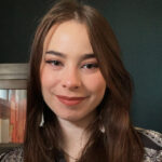 A portrait of a woman with long brown hair. She is standing in front of a dark green wall with a bookshelf in the background.