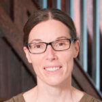 A portrait of a woman with glasses and brown hair pulled back, wearing a brown top. She is standing in front of a wooden and metal staircase.