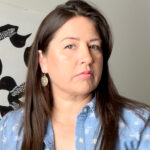 A woman with long brown hair wearing a light blue shirt with a white pattern and silver earrings. Behind her is a black and white patterned artwork featuring birds.