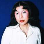 A woman with long black hair and pink eye makeup stands against a blue background. She is wearing a white button-up shirt and layered necklaces.