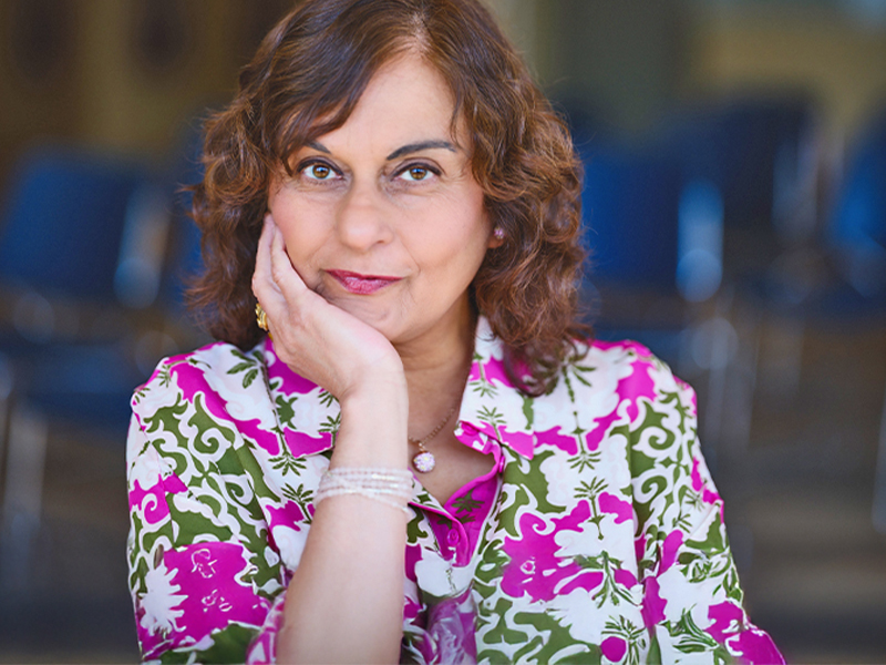 Kelly Kaur, with wavy shoulder-length brown hair and wearing a patterned white, green and pink top, poses with her chin resting on her hand.