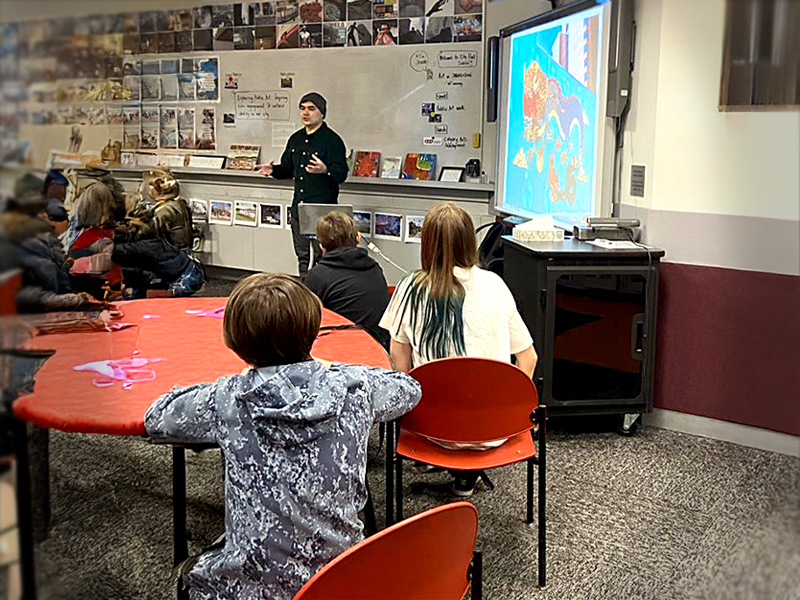 A teacher at the head of a classroom with a nearby screen showing his lesson as children listen