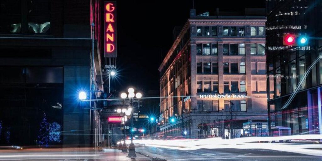 The Grand theatre at night with other buildings nearby, including the Downtown Hudson's Bay store. There are light trails from traffic on long exposure. The Grand's neon red sign shines out on the front of it, written vertically.