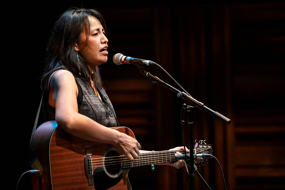 A singer with a guitar performs at a mic wearing beaded earrings and a vest, with long dark hair. Feels like its probably a previous artist from this Indigenous residency.