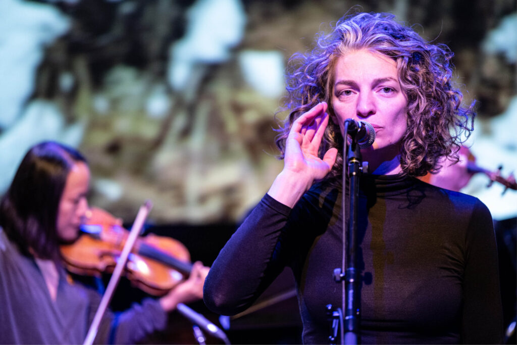 A singer with wavy shoulder length hair at a microphone performing. A violin player is slightly blurred in the background, also performing. The performers are lit by colourful studio lighting.
