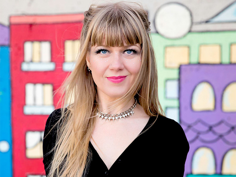 Tracy Wylie with long hair, wearing a black shirt standing against a wall with illustrated buildings on it