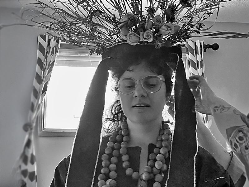Wednesday Lupypciw wearing glasses, looking downwards as hands adjust an elaborate floral/twig headpiece on their head