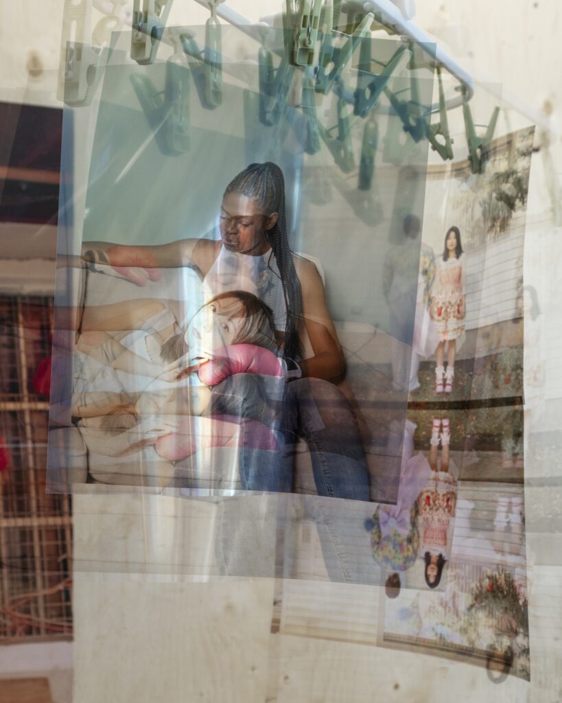 This is an artwork or maybe a photo of artwork, with layers and layers of reflection through glass and printed materials. It looks like a person with long black hair laying down on a the lap of another person with black braided hair looking over them and images are seen in the background of a duo with colourful clothes, and that is reflected underneath in a mirrored fashion.
