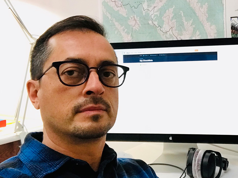 Bruno Canadien wearing dark-rimmed glasses and a blue and black plaid shirt sitting in front of a computer screen looking outwards